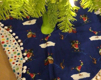 Puede incluir: Una falda circular azul para árbol de Navidad con un patrón repetitivo de un personaje verde con traje de Papá Noel rojo realizando actividades invernales. El borde está adornado con pompones blancos y el reverso muestra tela blanca con lunares de colores.