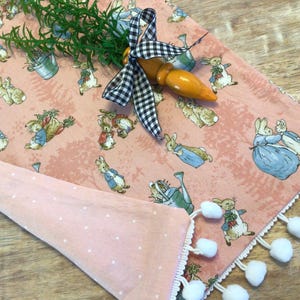 May include: A pink table runner with a white pom-pom trim and a Peter Rabbit print. The runner features a black and white checkered ribbon and a carrot.