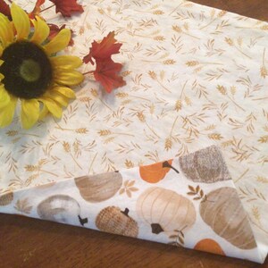 May include: Reversible fall-themed table runner. One side features a cream background with wheat stalks, the other side has pumpkins in shades of orange, brown, and cream. A yellow sunflower and fall leaves are placed on the runner.
