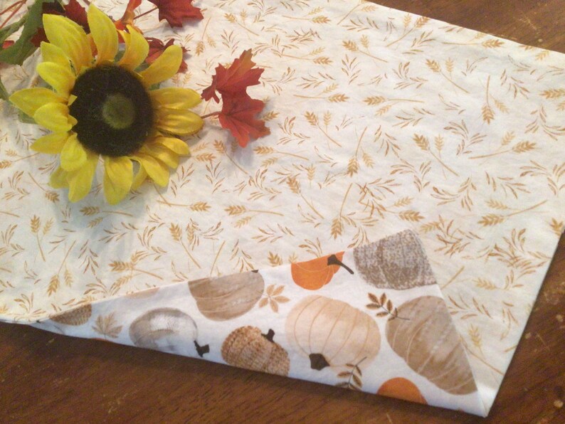 May include: Reversible fall-themed table runner. One side features a cream background with wheat stalks, the other side has pumpkins in shades of orange, brown, and cream. A yellow sunflower and fall leaves are placed on the runner.