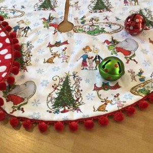 May include: A white Christmas tree skirt with a red pom-pom trim and a festive print of characters from the book "How the Grinch Stole Christmas". The skirt features two jingle bells, one red and one green.