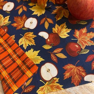 May include: A navy blue fabric with a fall theme, featuring red apples, apple slices, and autumn leaves in shades of yellow, orange, and brown. A section of orange plaid fabric is visible. Decorative orange flowers and a whole red apple are also present.