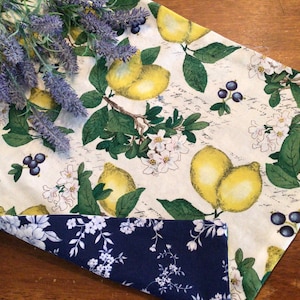 May include: A white table runner with a lemon and floral print. The runner has a blue and white floral fabric on the underside.