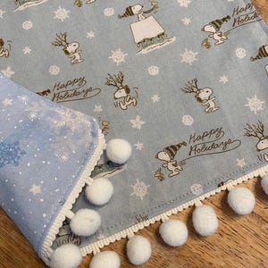 May include: A light blue holiday-themed table runner featuring Snoopy characters with reindeer antlers and the text "Happy Holidays." The runner has a white pom-pom trim and a snowflake pattern on the edge.