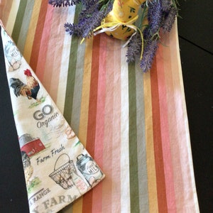 May include: A farmhouse-themed kitchen towel with a rooster illustration and the text "Go Organic Farm Fresh". The towel is folded next to a striped runner in shades of pink, green, and beige. A yellow fabric bunny and lavender sprigs are on top.