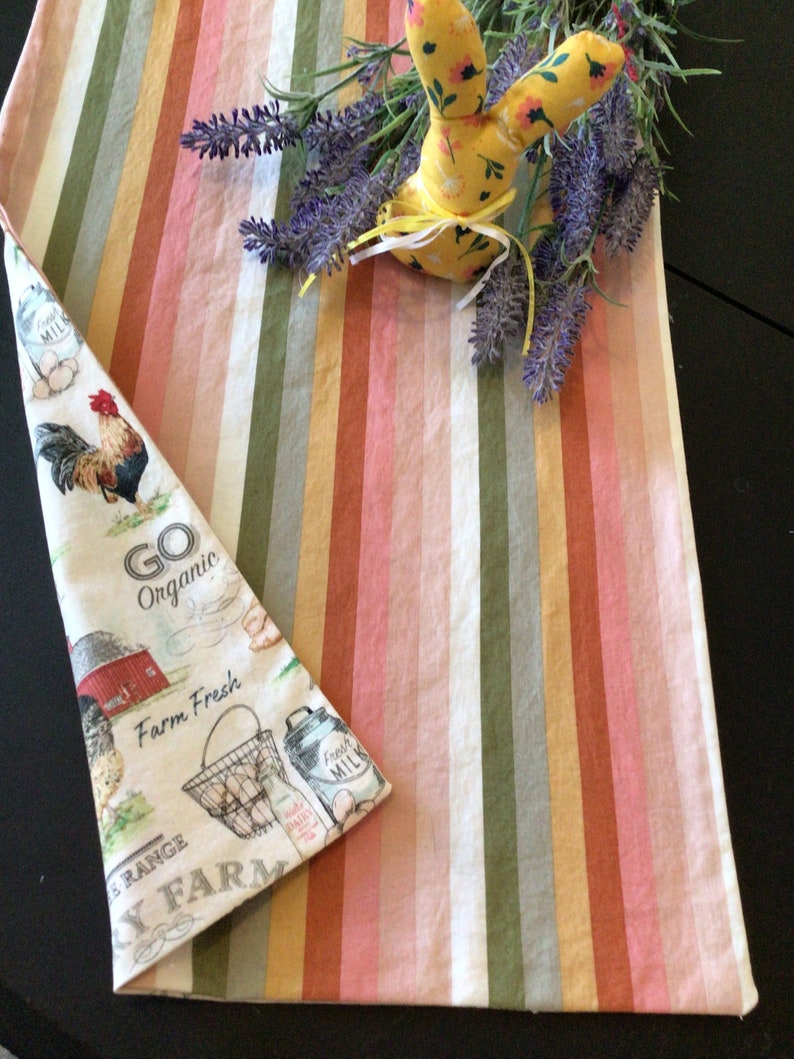 May include: A farmhouse-themed kitchen towel with a rooster illustration and the text "Go Organic Farm Fresh". The towel is folded next to a striped runner in shades of pink, green, and beige. A yellow fabric bunny and lavender sprigs are on top.