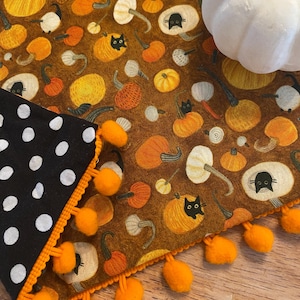 May include: A fabric table runner featuring a brown background with a pattern of orange pumpkins and black cats, accented by a black and white polka dot corner and orange pom-pom trim, with a white decorative pumpkin placed on top.