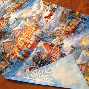 May include: A blue and white fabric table runner with a winter village scene. The runner features a snowy town with buildings, trees, and people. The fabric has a snowflake pattern on the back.