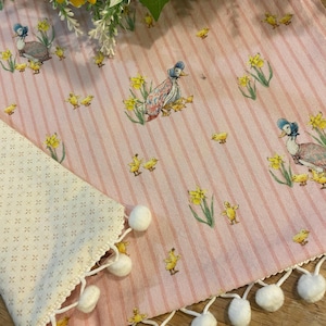 May include: Pink and white striped fabric with a duck and duckling pattern. The fabric has a white pom-pom trim. Yellow daffodils are also printed on the fabric. A section of off-white fabric with a small cross pattern is visible.