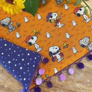 May include: A Halloween-themed bandana featuring Snoopy in various costumes, including a mummy and vampire, on an orange background with ghost and insect illustrations. The reverse side is dark blue with white stars. The edges are trimmed with purple pom-poms.