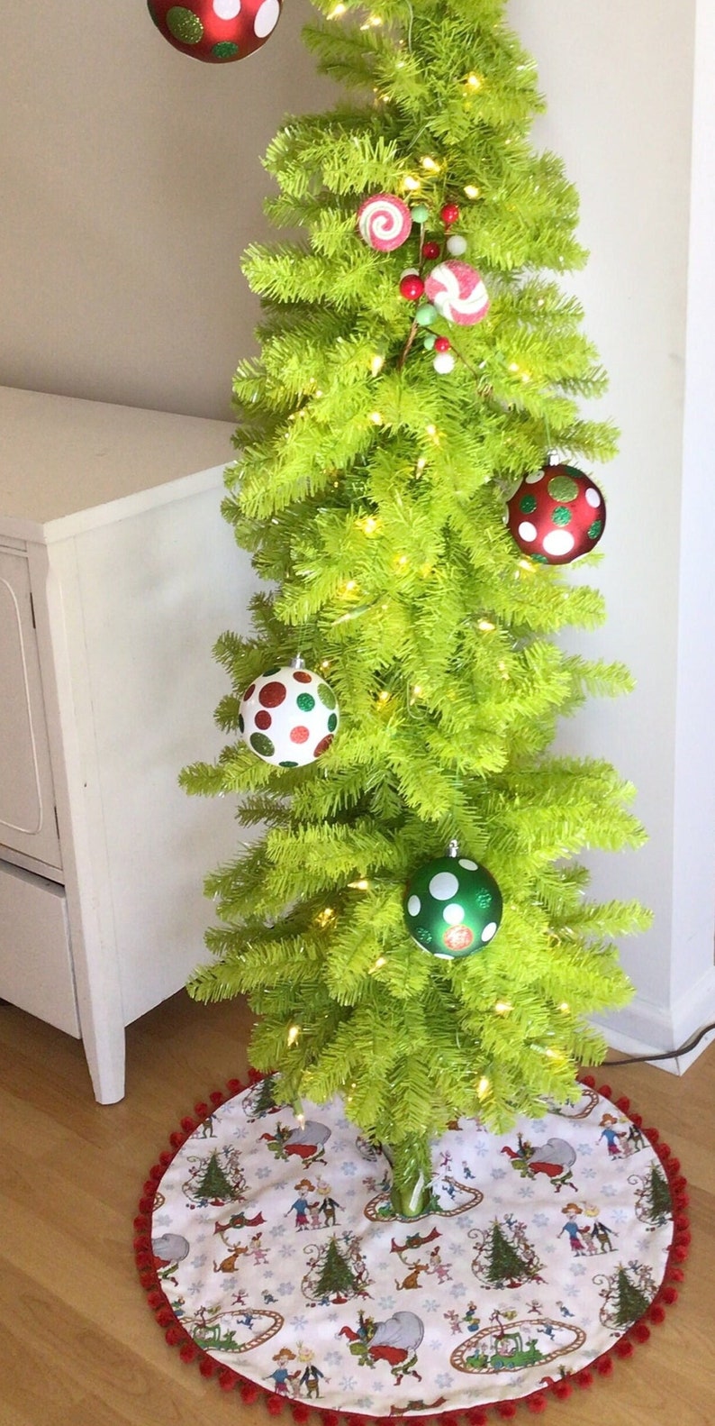 May include: A green artificial Christmas tree decorated with red and white polka dot ornaments, candy cane ornaments, and white lights. The tree is sitting on a white and red tree skirt with a whimsical Christmas scene.