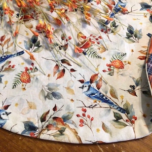 May include: A white fabric table runner with a blue jay, leaf, and berry pattern. The edges are trimmed with a plaid fabric in shades of orange, blue, and white.