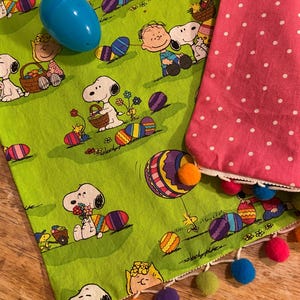 May include: A green fabric featuring Snoopy and friends with Easter eggs and baskets. A blue plastic egg sits on top. A pink polka dot fabric with a white lace trim and colorful pom-poms is also visible.