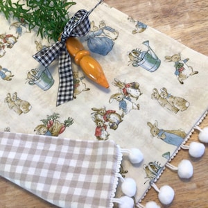 May include: A white table runner with a gingham border and a white pom-pom trim. The runner features a repeating pattern of bunnies and gardening tools. A carrot-shaped wooden ornament is placed on top of the runner.