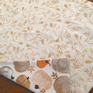 May include: Reversible fall-themed table runner. One side features a cream background with gold wheat stalks. The other side has a white background with pumpkins in shades of orange, brown, and gray. Perfect for autumn decor.