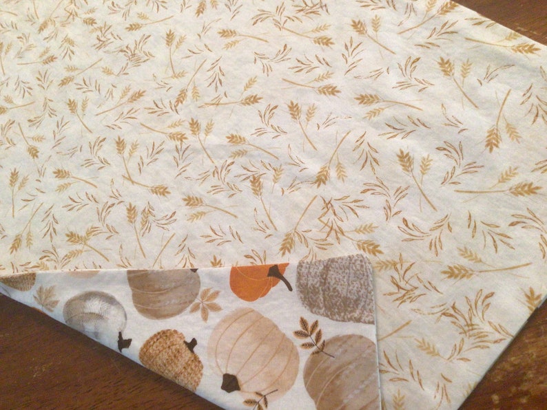 May include: Reversible fall-themed table runner. One side features a cream background with gold wheat stalks. The other side has a white background with pumpkins in shades of orange, brown, and gray. Perfect for autumn decor.