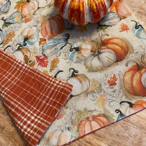 May include: A fall-themed table runner with a pumpkin and a plaid napkin. The runner features a pattern of pumpkins in shades of orange, blue, and white, with autumn leaves on a cream background. The napkin is orange and white plaid.