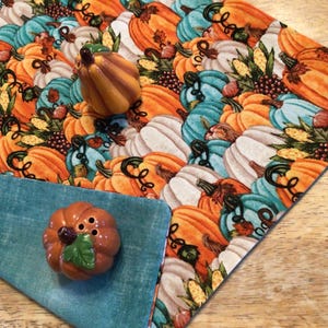 May include: A fall-themed table runner with a pumpkin pattern, featuring orange, teal, and white pumpkins. Two pumpkin-shaped ceramic salt and pepper shakers sit on a teal napkin. The runner is on a wooden surface.