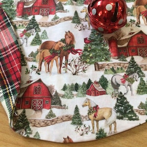 May include: Christmas tree skirt with a festive farm scene featuring red barns, horses, and evergreen trees on a snowy background. The skirt has a plaid border in red, green, and cream. A red jingle bell is in the image.