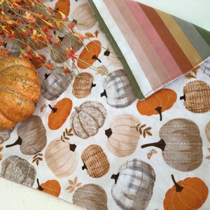 May include: A white table runner with a repeating pattern of pumpkins in shades of orange, tan, and gray. A large orange pumpkin and fall foliage are on the left. The runner is reversible with a striped pattern on the other side.