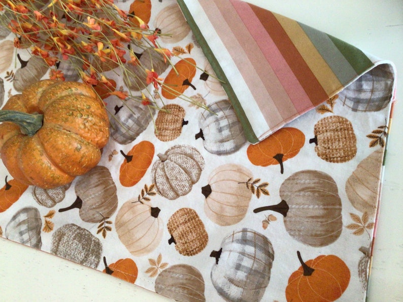 May include: A white table runner with a repeating pattern of pumpkins in shades of orange, tan, and gray. A large orange pumpkin and fall foliage are on the left. The runner is reversible with a striped pattern on the other side.