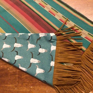 May include: A turquoise table runner with a white skull pattern and brown fringe. The runner has a striped pattern on the back side.