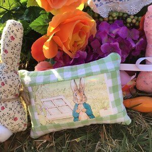 May include: A green and white checkered pillow with a picture of Peter Rabbit in a blue jacket. The pillow is surrounded by artificial flowers, including pink roses, orange roses, and purple hydrangeas. There are also two fabric bunny toys, one pink and one cream.