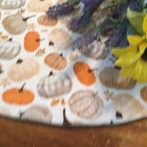 May include: A round table runner with a repeating pattern of pumpkins in shades of orange, tan, and white on a white background. The runner is decorated with a sunflower and lavender sprigs.