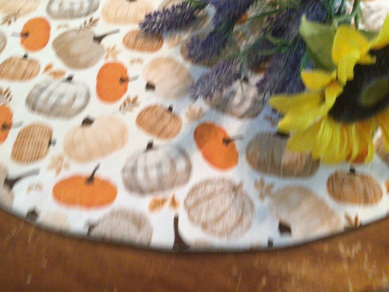 May include: A round table runner with a repeating pattern of pumpkins in shades of orange, tan, and white on a white background. The runner is decorated with a sunflower and lavender sprigs.