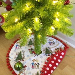 May include: A Christmas tree skirt with a whimsical design featuring characters and trees. The skirt is white with a red polka dot border and red pom-pom trim. A green bell and a Christmas tree are also visible.
