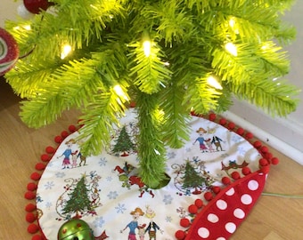 May include: A Christmas tree skirt with a whimsical design featuring characters and trees. The skirt is white with a red polka dot border and red pom-pom trim. A green bell and a Christmas tree are also visible.