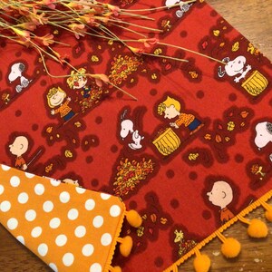 May include: Red and orange fabric table runner with a white polka dot pattern on the reverse side. The runner features a repeating pattern of Peanuts characters, including Snoopy, Charlie Brown, and Lucy, with autumn leaves. The runner has a yellow pom-pom trim.