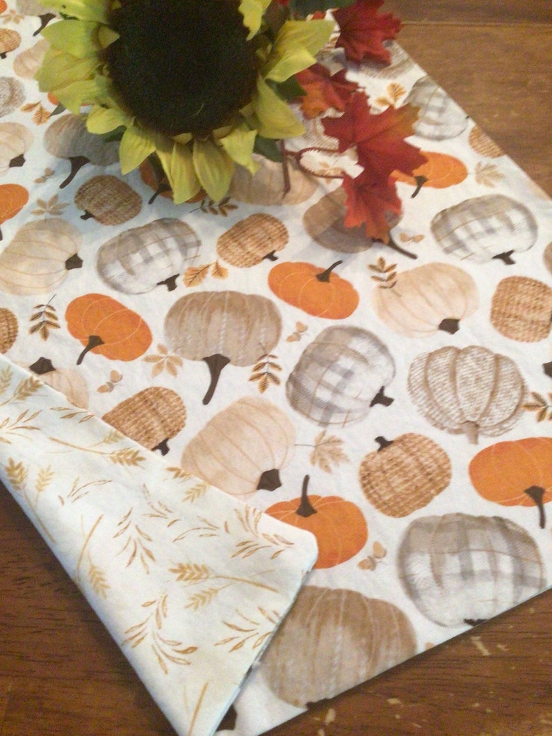 May include: A reversible fall-themed table runner featuring a pumpkin pattern in orange, tan, and gray on a white background. The reverse side has a wheat stalk design. Artificial sunflowers and fall leaves are placed on the runner.