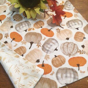 May include: A white table runner with a repeating pattern of pumpkins in shades of orange, beige, and gray. The runner is decorated with a sunflower and fall leaves. A folded napkin with a wheat pattern is also visible.