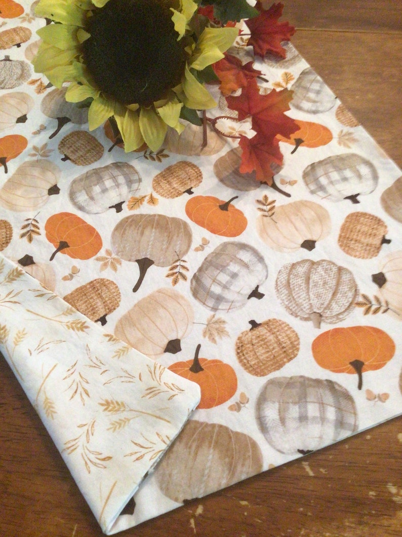 May include: A white table runner with a repeating pattern of pumpkins in shades of orange, beige, and gray. The runner is decorated with a sunflower and fall leaves. A folded napkin with a wheat pattern is also visible.