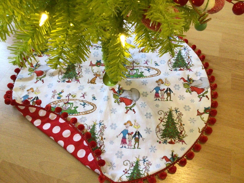May include: A white Christmas tree skirt with a red and white polka dot trim and red pom-poms around the edge. The skirt features a whimsical illustration of characters from the book "The Grinch" by Dr. Seuss.