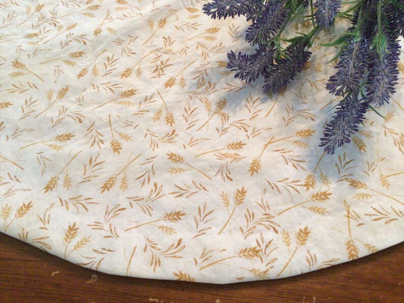 May include: A cream-colored fabric with a repeating pattern of wheat stalks in shades of gold and brown. The fabric is slightly curved, suggesting it is part of a larger piece. A sprig of purple lavender is in the upper right corner.