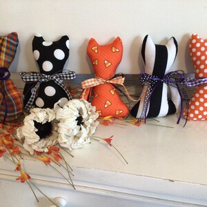 May include: A collection of handmade cat-shaped fabric dolls in various Halloween-themed patterns. The dolls feature patterns like plaid, polka dots, candy corn, and stripes, each tied with a decorative ribbon. White flowers and orange floral accents are also present.