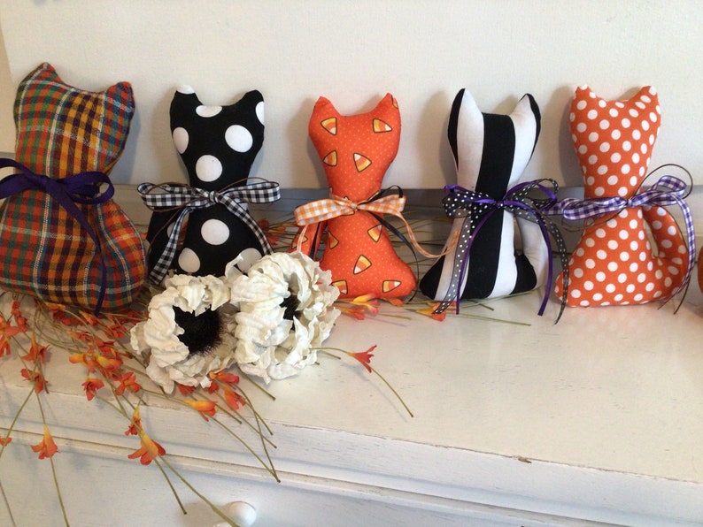 May include: A collection of handmade cat-shaped fabric dolls in various Halloween-themed patterns. The dolls feature patterns like plaid, polka dots, candy corn, and stripes, each tied with a decorative ribbon. White flowers and orange floral accents are also present.