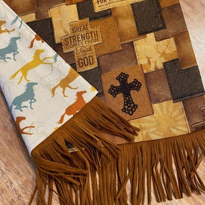 May include: A brown and tan fabric scarf with a fringe border. The scarf features a patchwork design with various inspirational quotes and images, including a cross, horses, and the words "Great Strength Comes From Faith in God".