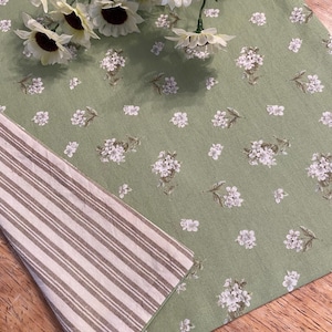 May include: A green floral table runner with white flower and leaf patterns. The runner is paired with a striped fabric. Artificial sunflowers are placed on top of the runner. The fabrics are on a wooden surface.