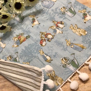 May include: A blue and white table runner with a Peter Rabbit design. The runner has a white fringe with white pom-poms along the edges. The runner is made of cotton and is perfect for a spring or summer table setting.