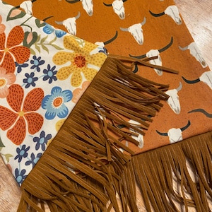 May include: A brown and white fabric scarf with a floral pattern on one side and a pattern of white cow skulls on a brown background on the other side. The scarf has brown fringe on both ends.