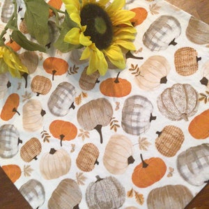 May include: A white table runner with a repeating pattern of pumpkins in orange, tan, and gray. Some pumpkins have plaid or woven textures. Yellow sunflowers are placed on the runner.