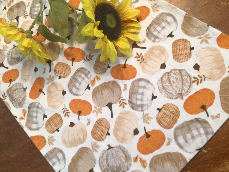 May include: A white table runner with a repeating pattern of pumpkins in orange, tan, and gray. Some pumpkins have plaid or woven textures. Yellow sunflowers are placed on the runner.