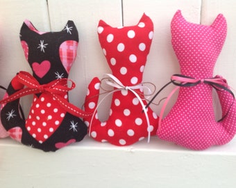 May include: Four handmade fabric cat toys. The cats are in various colors and patterns, including red, pink, black, and white polka dots. Each cat has a ribbon tied around its neck.