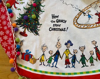 May include: A white Christmas tree skirt with a red and white border and pom-poms. The skirt features a print of characters from the book "How the Grinch Stole Christmas!" The text "How the Grinch Stole Christmas!" is printed on the skirt.
