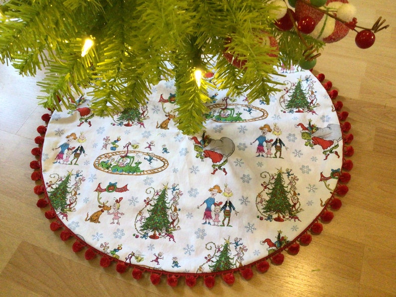 May include: A white Christmas tree skirt with a red pom-pom trim. The skirt features a whimsical illustration of characters from the classic children's book "How the Grinch Stole Christmas!"