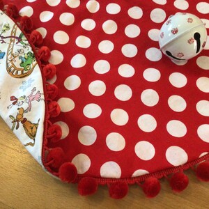 May include: Red and white polka dot Christmas tree skirt with a red pom-pom trim. The skirt is folded over to show a white fabric with a whimsical Christmas scene.