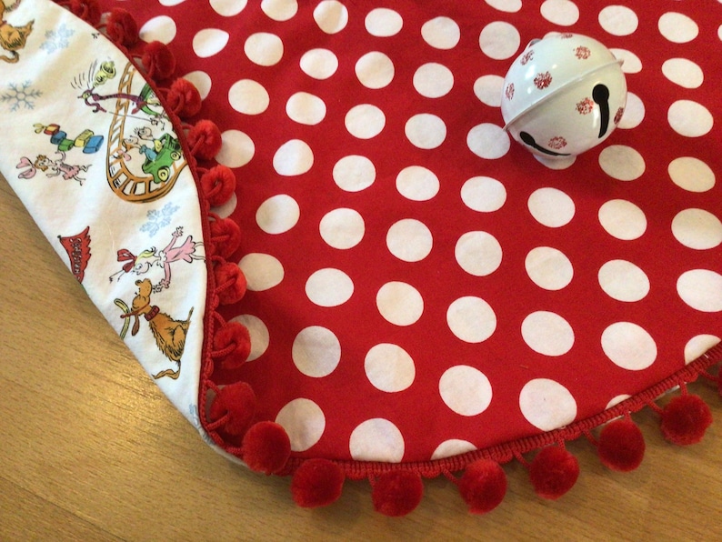 May include: Red and white polka dot Christmas tree skirt with a red pom-pom trim. The skirt is folded over to show a white fabric with a whimsical Christmas scene.